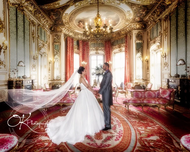Bride and groom holding hands in a luxurious, ornate room with red and gold decor and a grand chandelier.