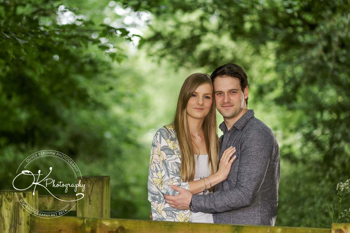 A couple posing together with arms around each other, standing in front of a lush, green forest background.