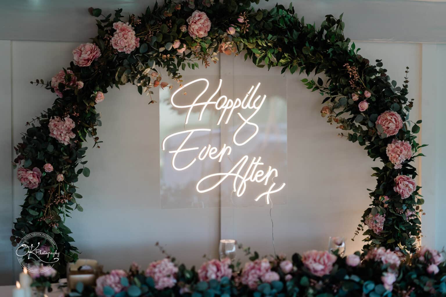 A beautifully decorated arch featuring greenery and pink flowers, with a glowing neon sign that reads "Happily Ever After" in elegant cursive lettering.
