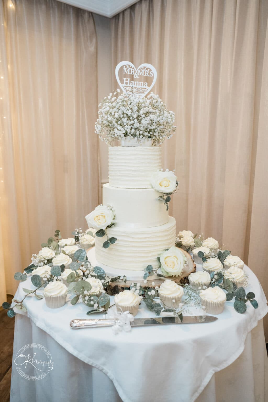 Three-tier wedding cake with white frosting, decorated with white roses, greenery, and topped with baby's breath and a "Mr & Mrs Hanna" heart-shaped topper. The cake table also has matching cupcakes and a cake knife resting on a white tablecloth.