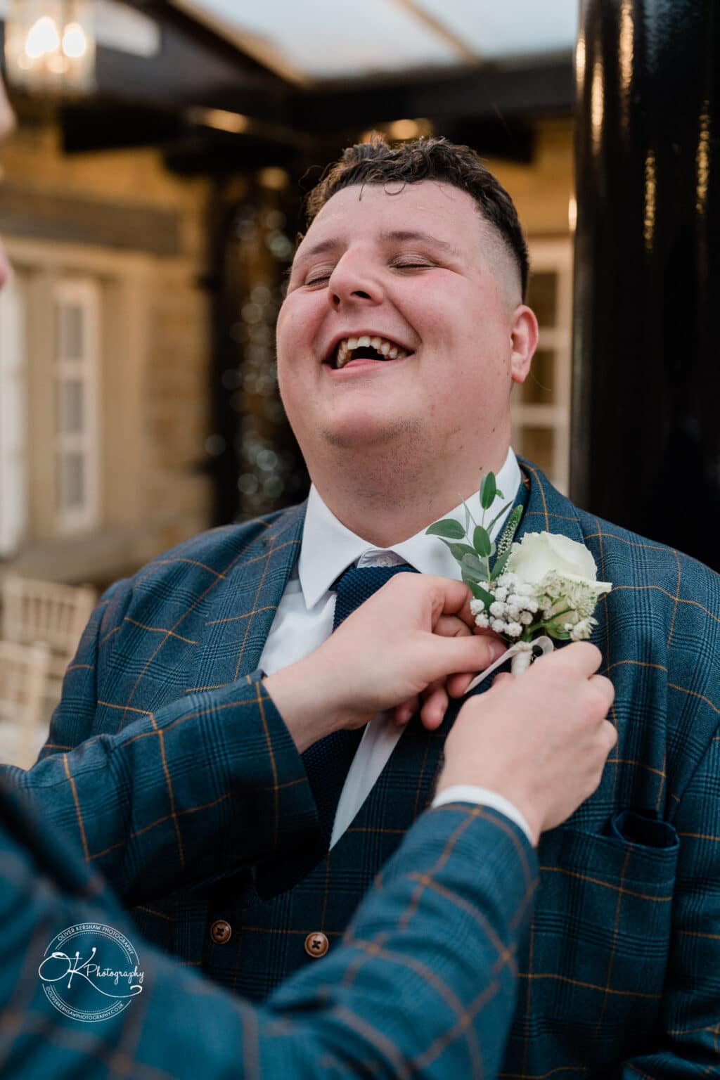 A man wearing a checkered suit and tie smiles while another person adjusts his boutonniere.