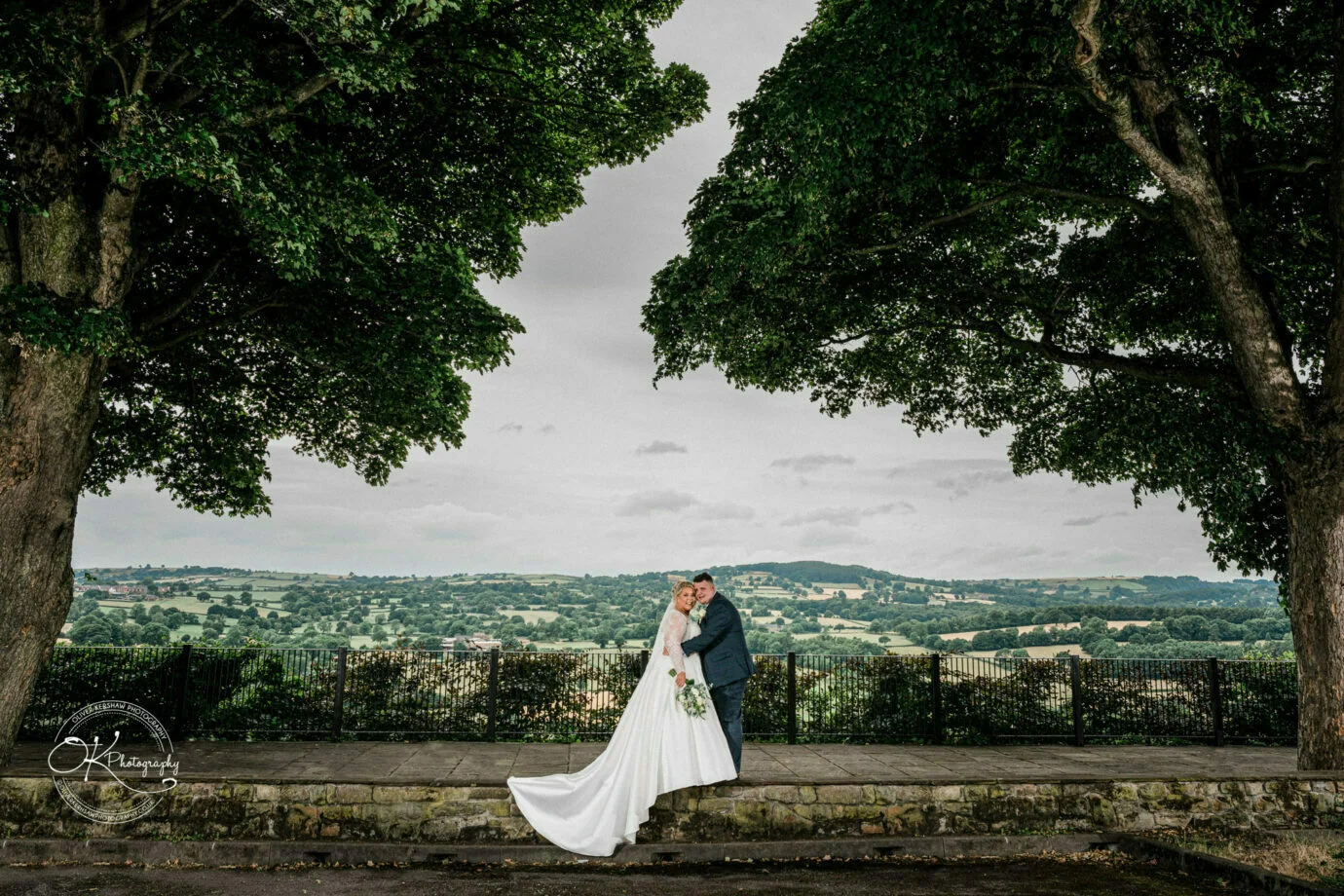 A wedding couple embracing under two large trees with a scenic countryside in the background.