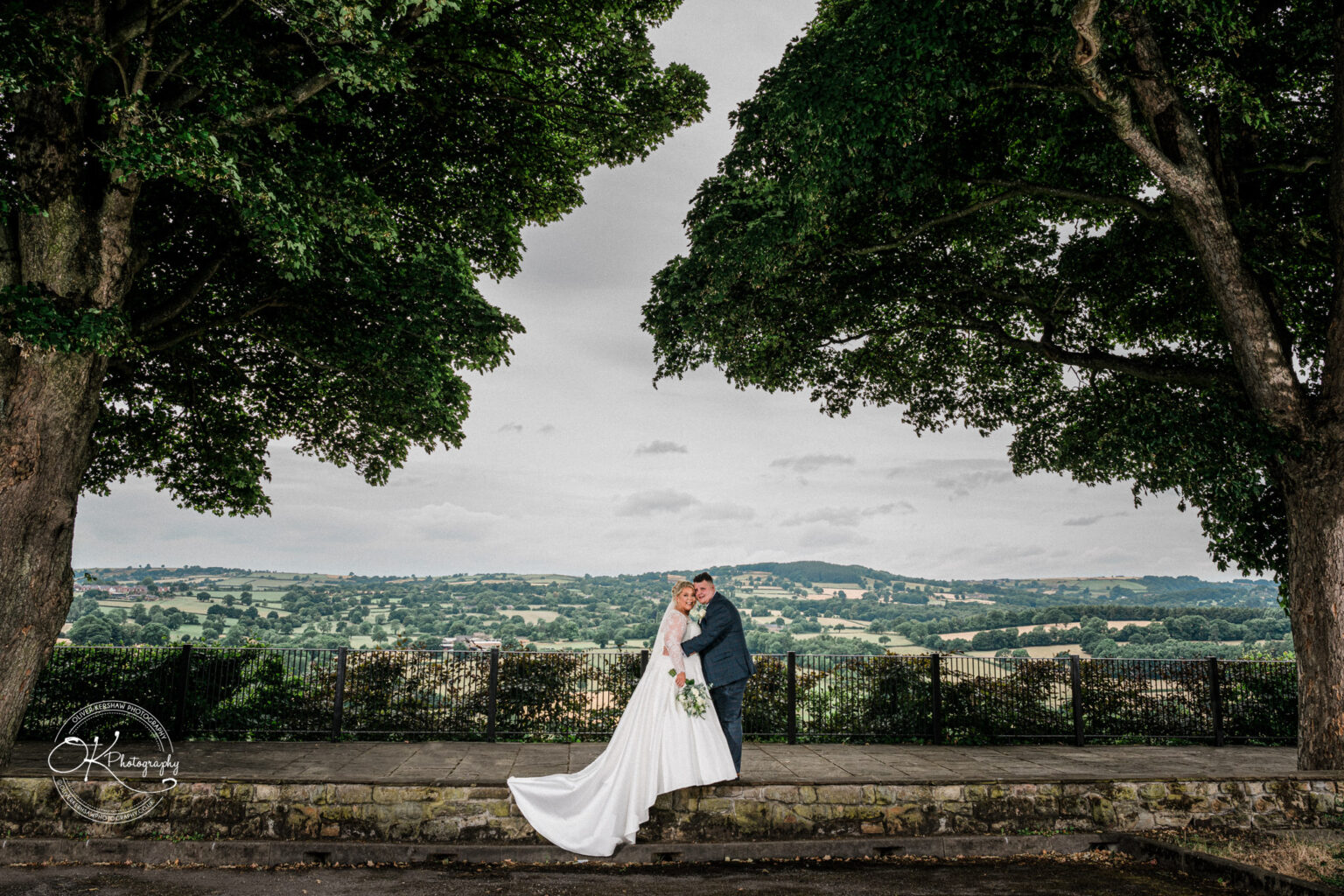 Santos Higham Farm Wedding Photography A wedding couple embracing under two large trees with a scenic countryside in the background.