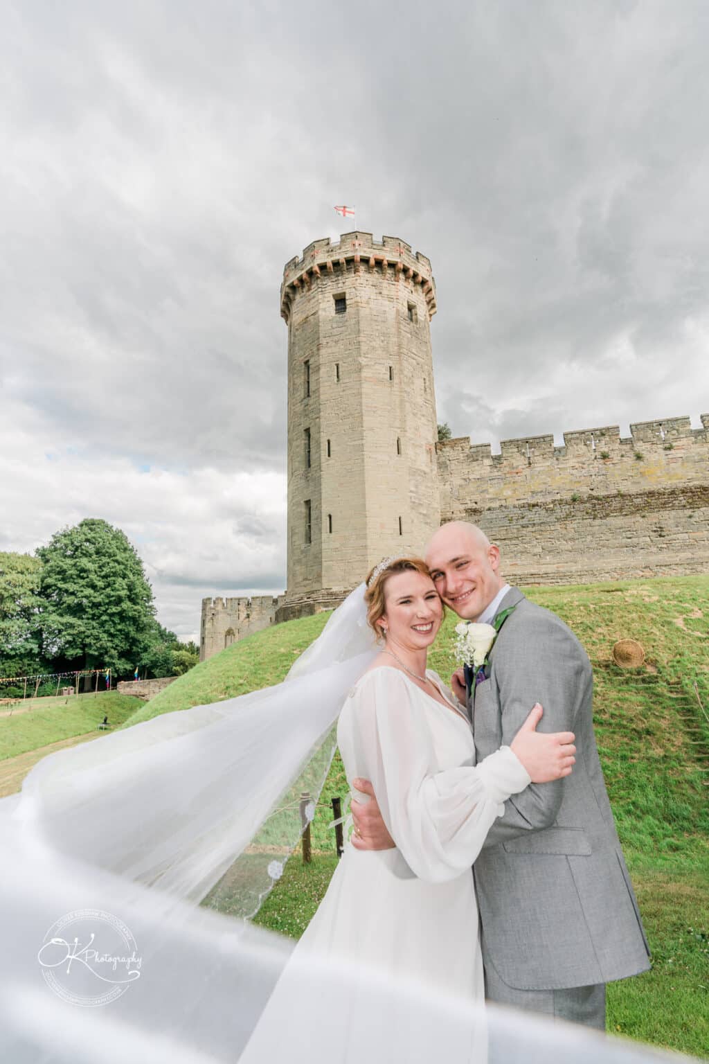 Bride and groom embracing in front of a historic stone castle with a turret and the flag of England on top, under an overcast sky.