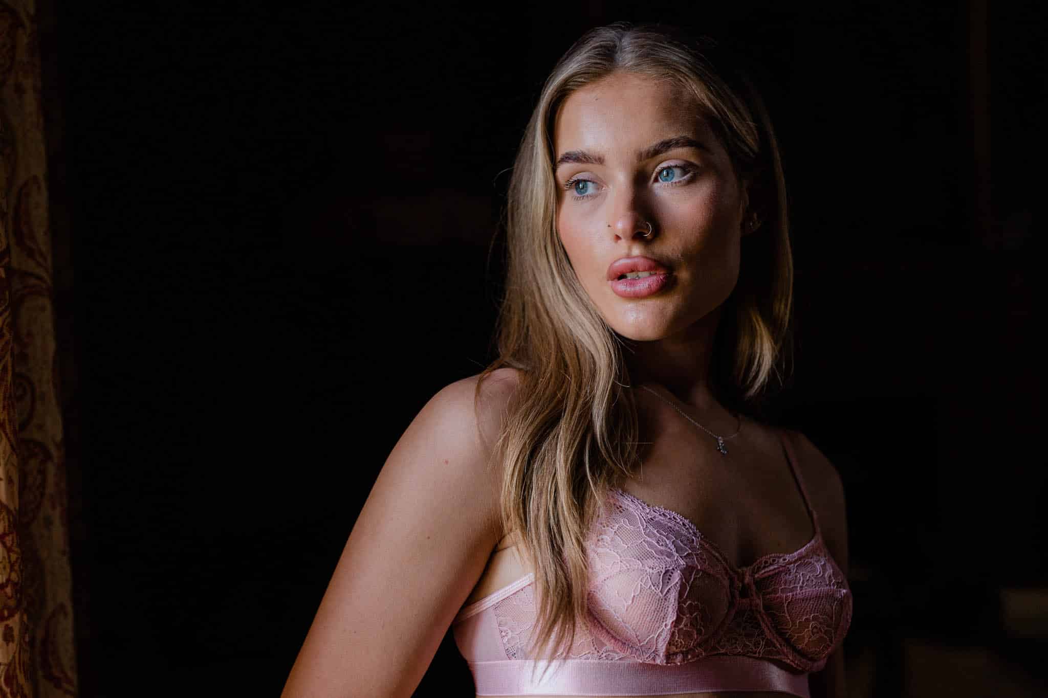 Woman with long blonde hair wearing a pink lace bralette, looking to the side with a dark background.