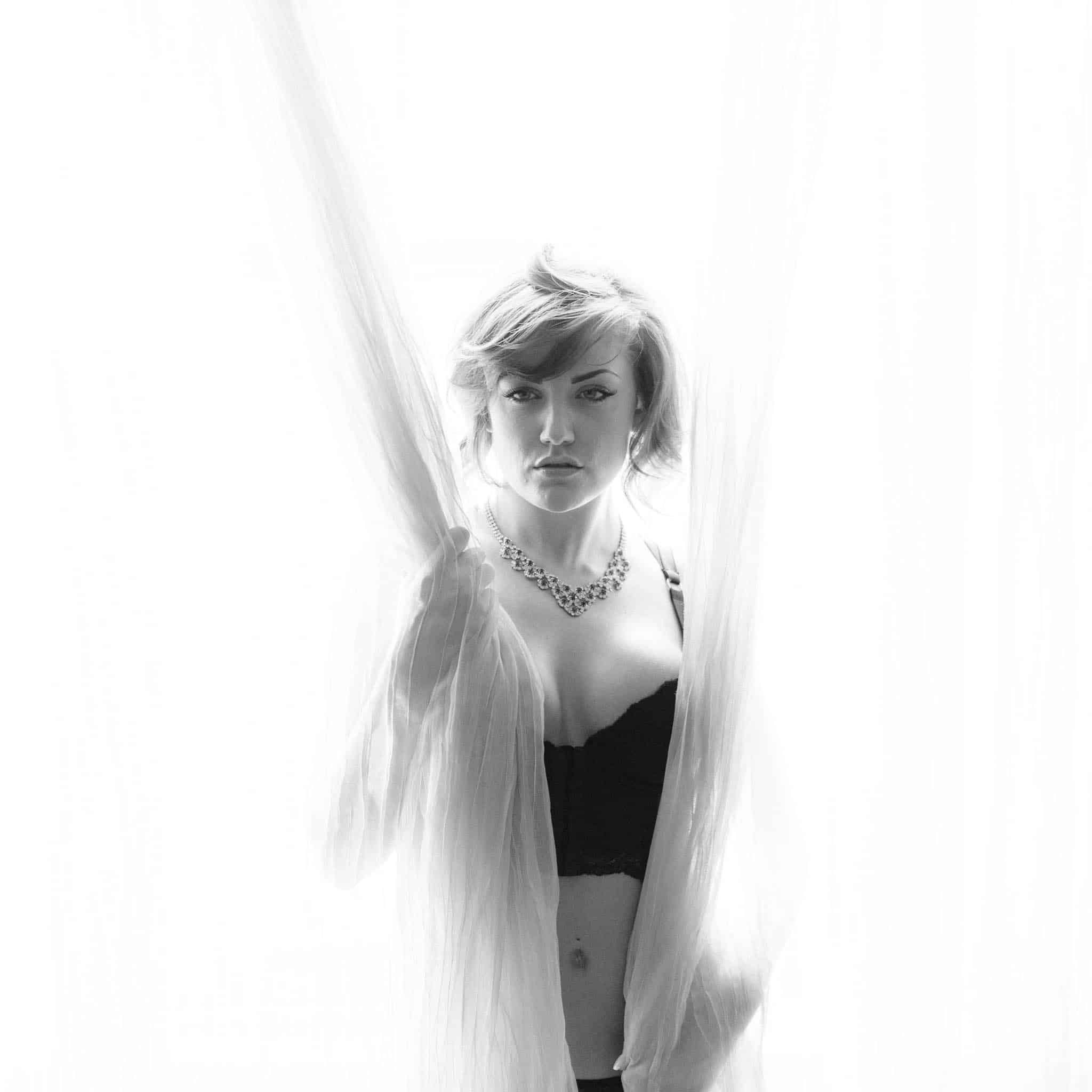 A woman in black lingerie stands behind sheer curtains in a high-key black and white photograph.
