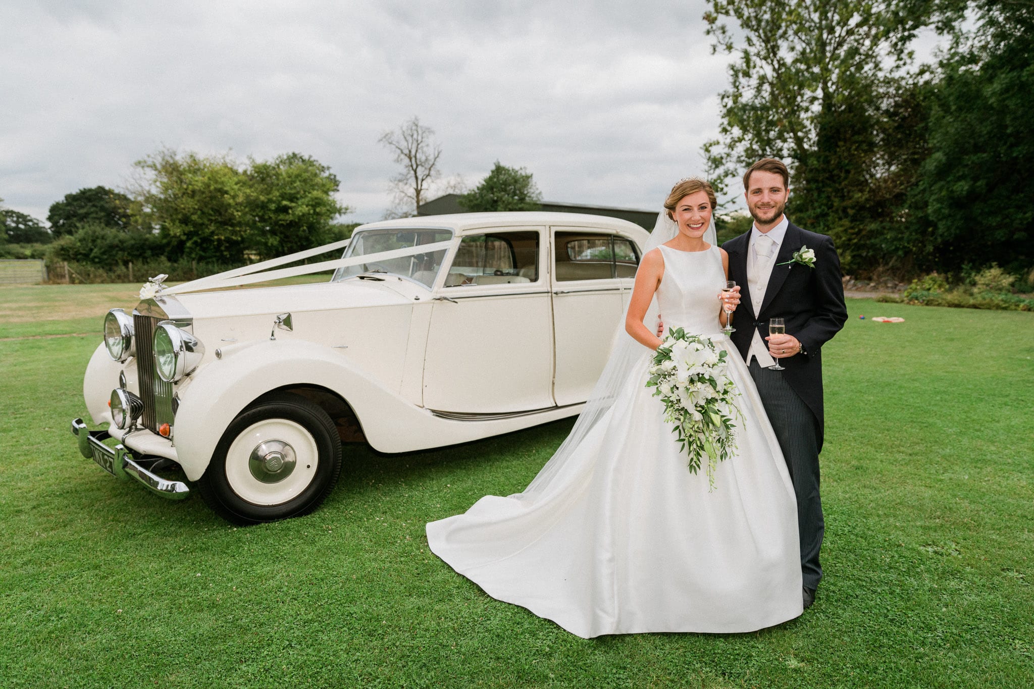 A bride and groom stand in front of a classic car at The Old Stables Wedding Venue in Leicestershire. The vintage and romantic setting creates a beautiful atmosphere for the couple to enjoy their special moment together.