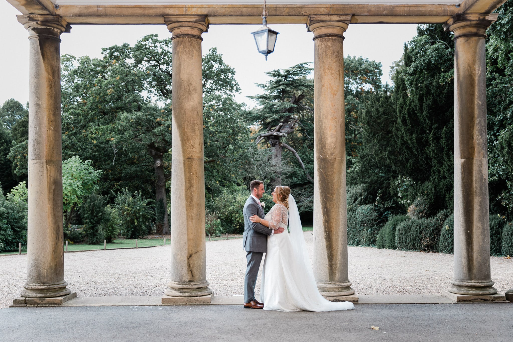 This image showcases wedding photography at Prestwold Hall, a stunning venue with impressive columns and beautiful architecture. The photo shows the couple standing in front of the columns, with the photographer expertly capturing the details of the venue and the love and joy of the couple on their wedding day. The image is beautifully lit, with the columns and architecture of the venue serving as a stunning backdrop for the couple's special day.