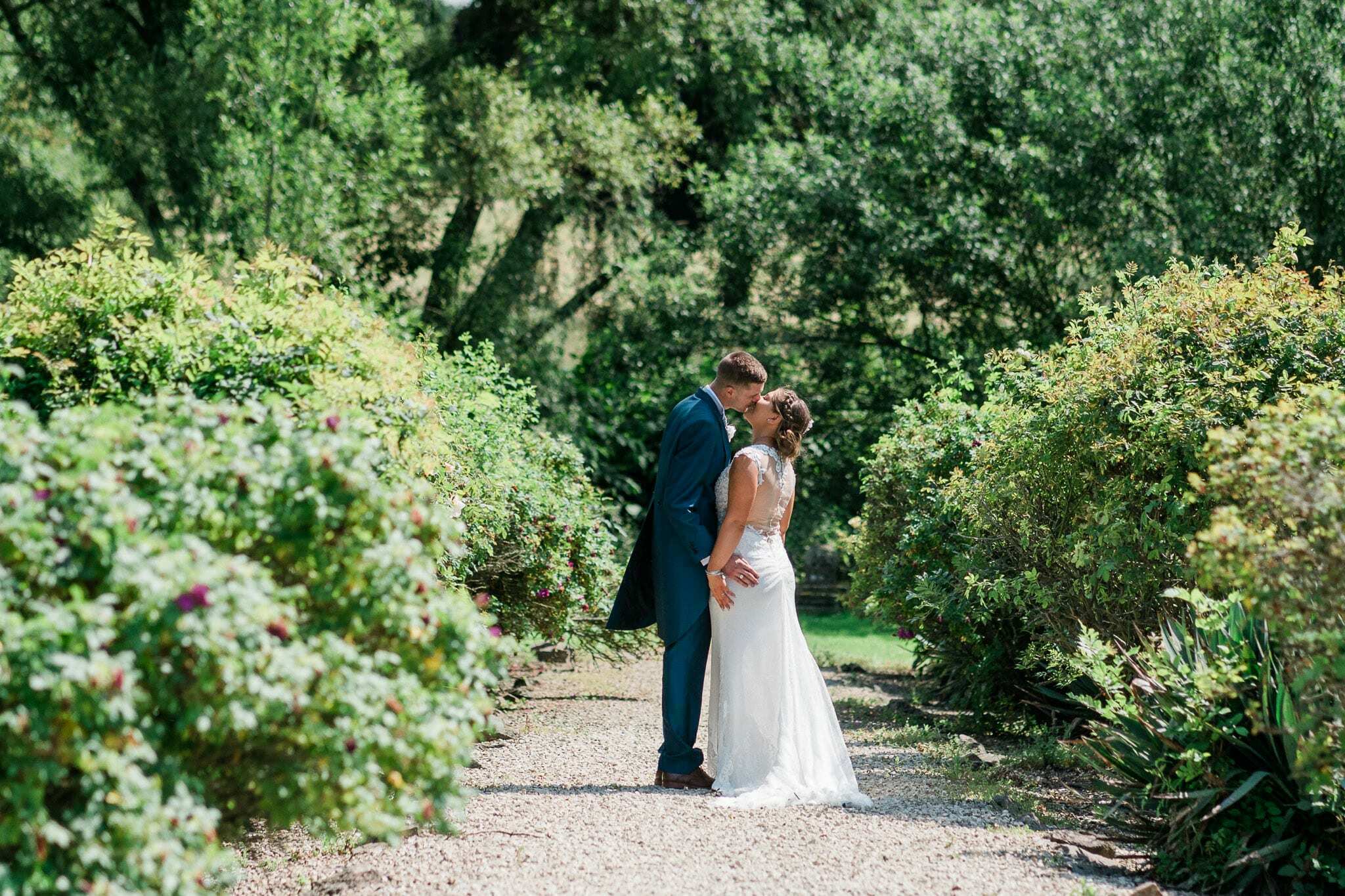 A bride and groom stand together in a garden, with the Rothley Court Hotel in the background. The bride wears a white gown and holds a bouquet of white flowers, while the groom is dressed in a tuxedo.