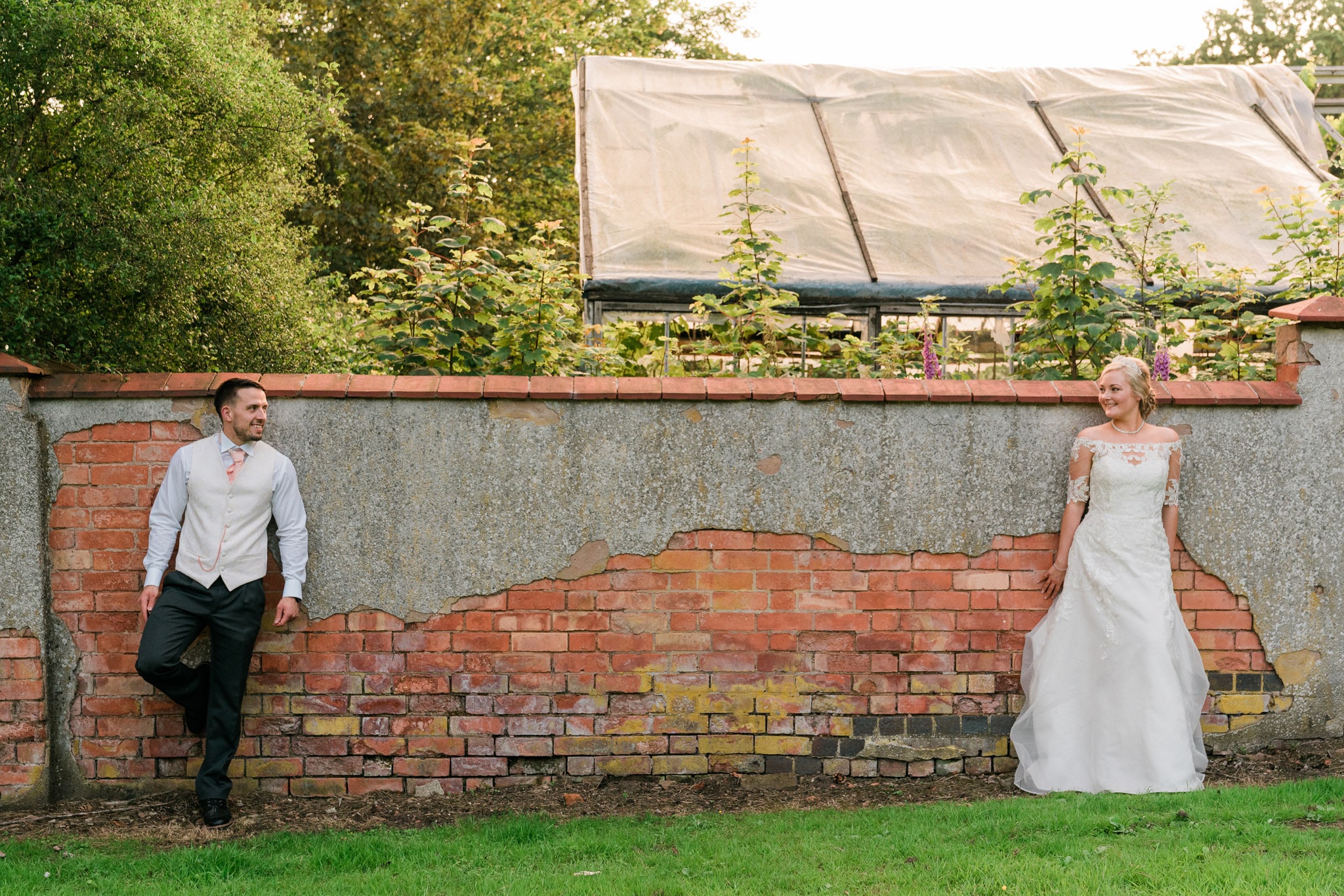 A bride and groom stand against a wall at Rothley Court Hotel, with an archway in the background. The bride wears a white lace dress with a long train, while the groom is dressed in a black tuxedo.
