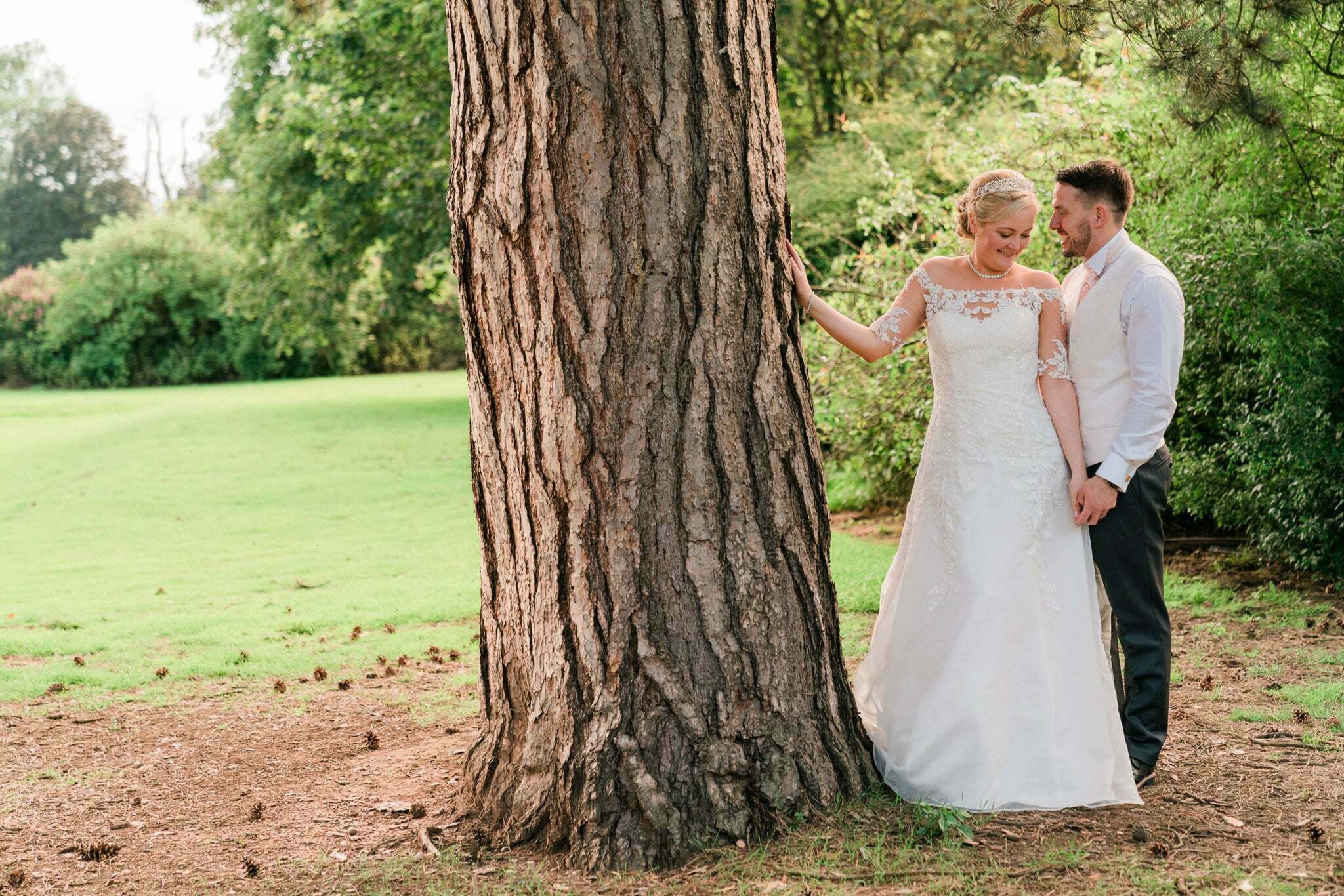 A bride and groom walk hand in hand in a rose garden at Rothley Court Hotel. The groom is wearing a black tuxedo while the bride is wearing a white lace dress with a long train. They smile at each other as they walk through the beautiful garden filled with colorful roses.