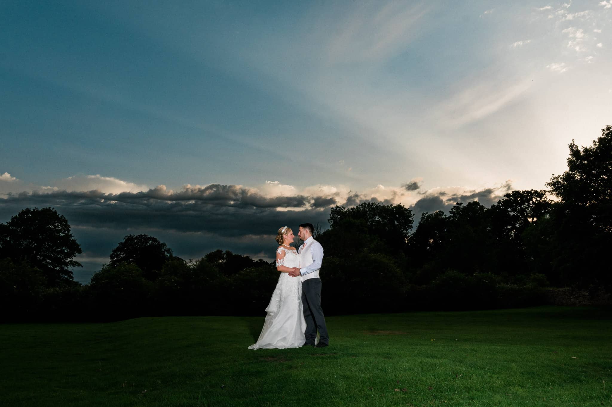 A bride and groom embrace in front of a stunning sunset at Rothley Court Hotel. The warm glow of the sunset casts a beautiful light on the couple, creating a romantic atmosphere.