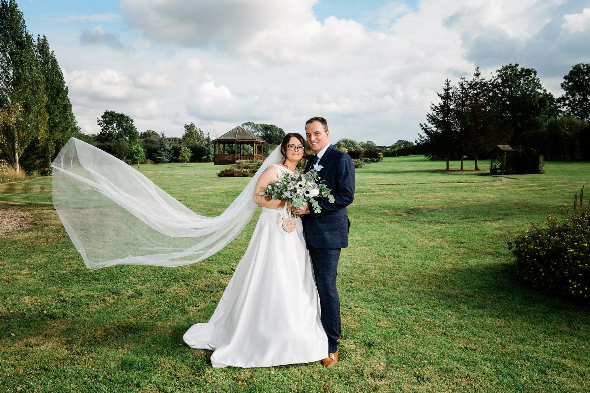 A beautiful bride and handsome groom stand in the gardens of Shearsby Bath wedding venue in Leicester. The bride's veil blows gently in the wind as they share a romantic moment captured expertly by photographer Oliver Kershaw. The natural setting and soft lighting of the gardens make the image even more enchanting, highlighting the beauty and wonder of wedding photography.