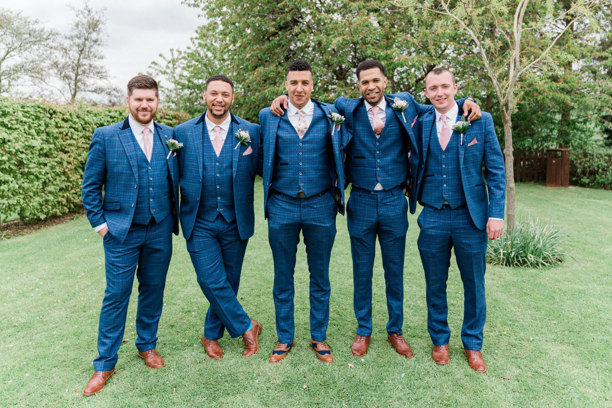 A group of handsome groomsmen stand together in the stunning grounds of Shearsby Bath wedding venue in Leicester. The lush greenery and charming architecture of the venue make for a beautiful backdrop as the men enjoy a moment of camaraderie captured expertly by photographer Oliver Kershaw. The dynamic and playful formation of the group adds to the visual appeal of the image, showcasing the fun and lightheartedness of the wedding party.