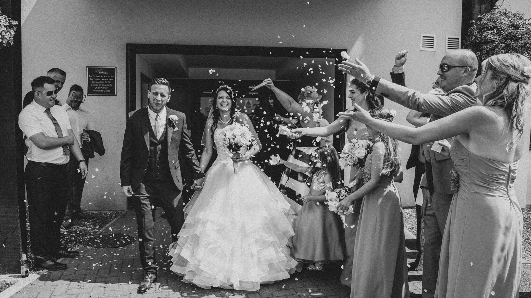 A bride and groom walk through a shower of confetti at Grange Hotel, surrounded by their wedding guests. The bride's white dress and veil contrast against the colorful confetti, and the groom smiles broadly as he looks at his bride.