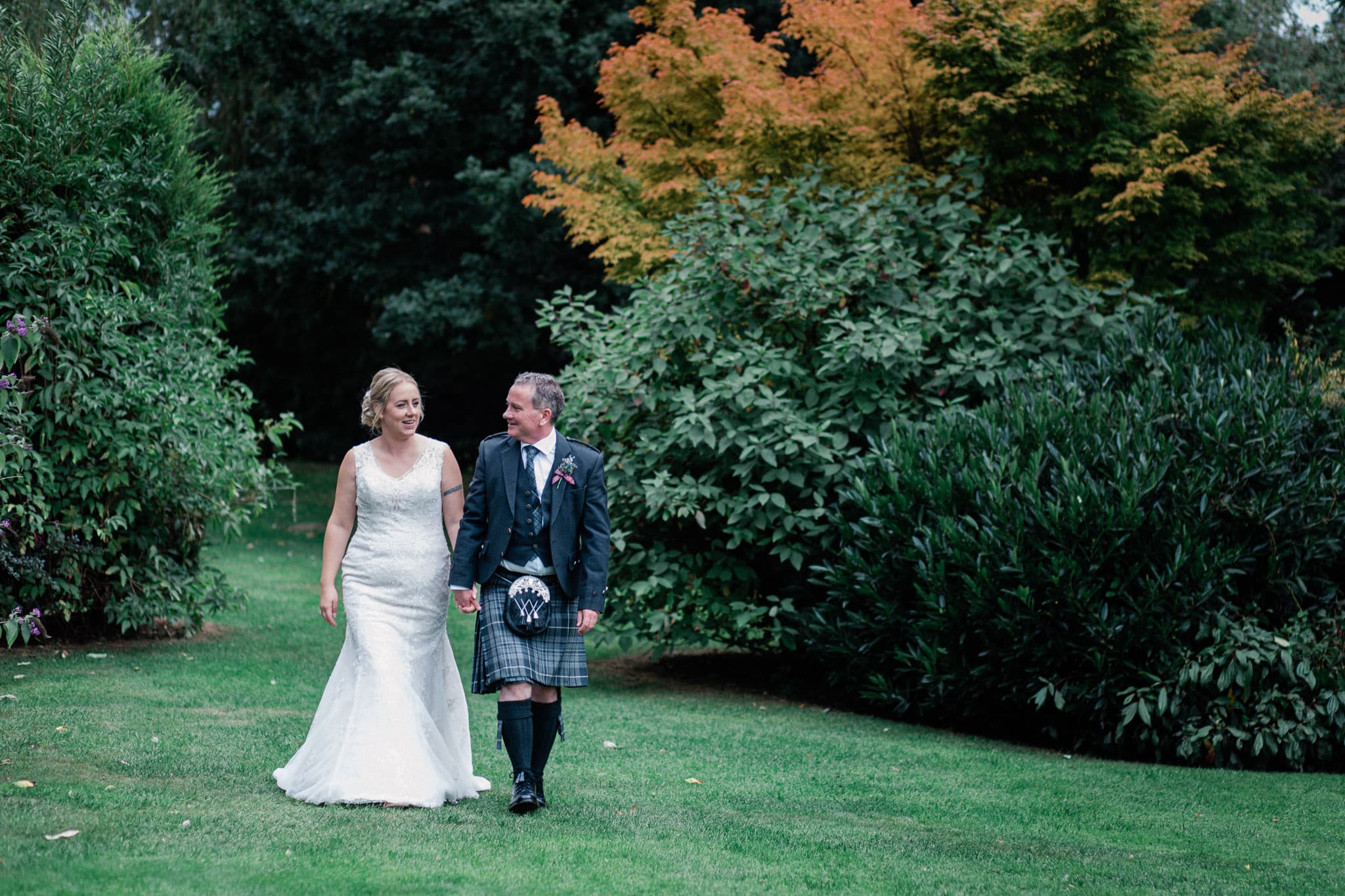A bride and groom walk hand in hand in the gardens of Sketchley Grange Hotel surrounded by blooming flowers and tall trees.