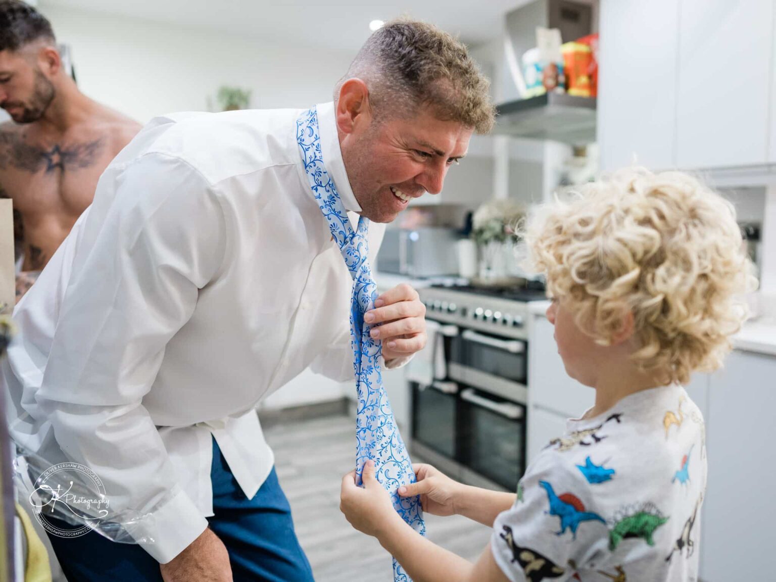 Man in a white shirt and blue trousers bending towards a boy with blonde curly hair, who is helping him with a tie in a kitchen.