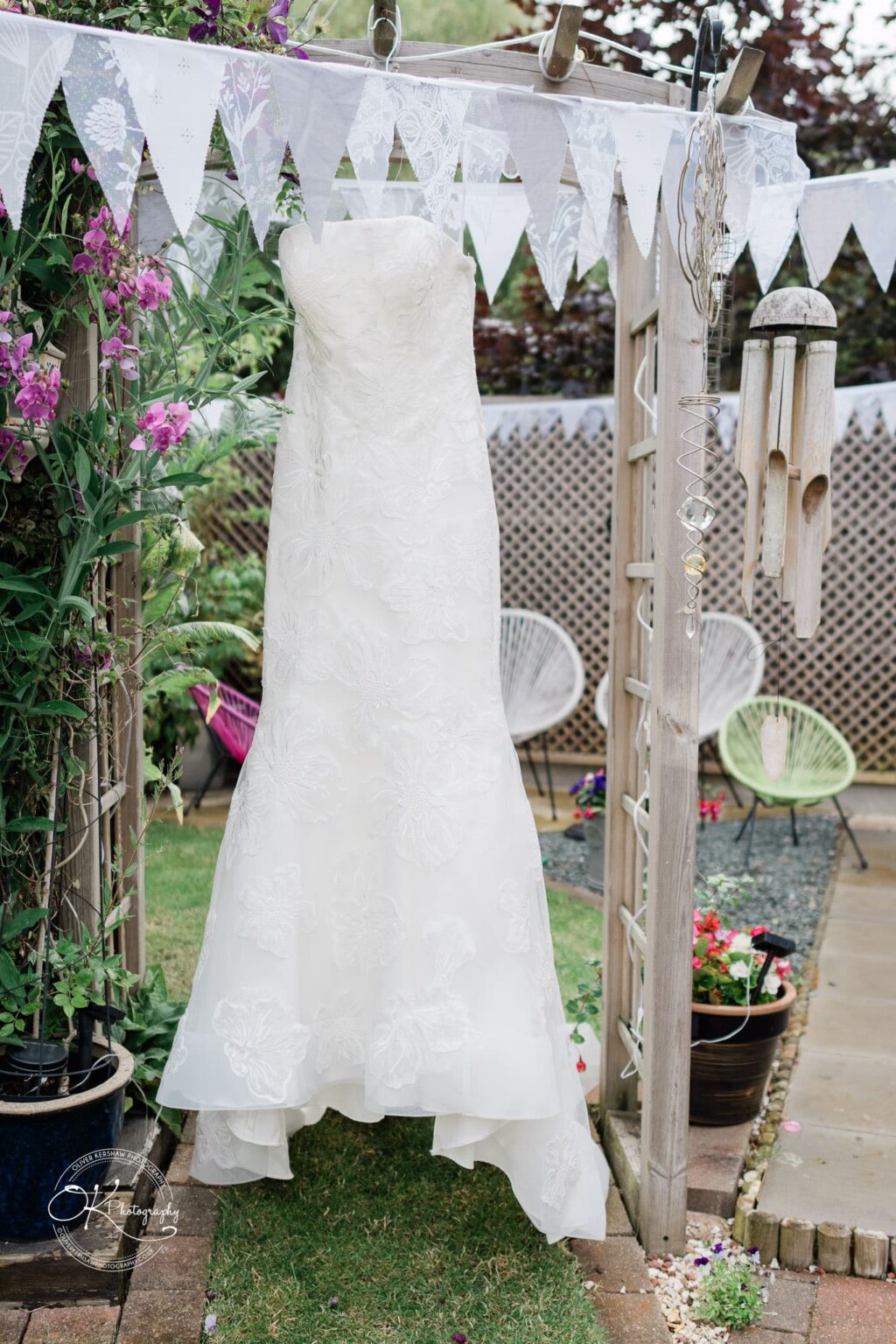 Strapless white wedding dress with floral lace details hanging on a wooden pergola decorated with white triangular bunting, surrounded by garden plants and a wind chime.