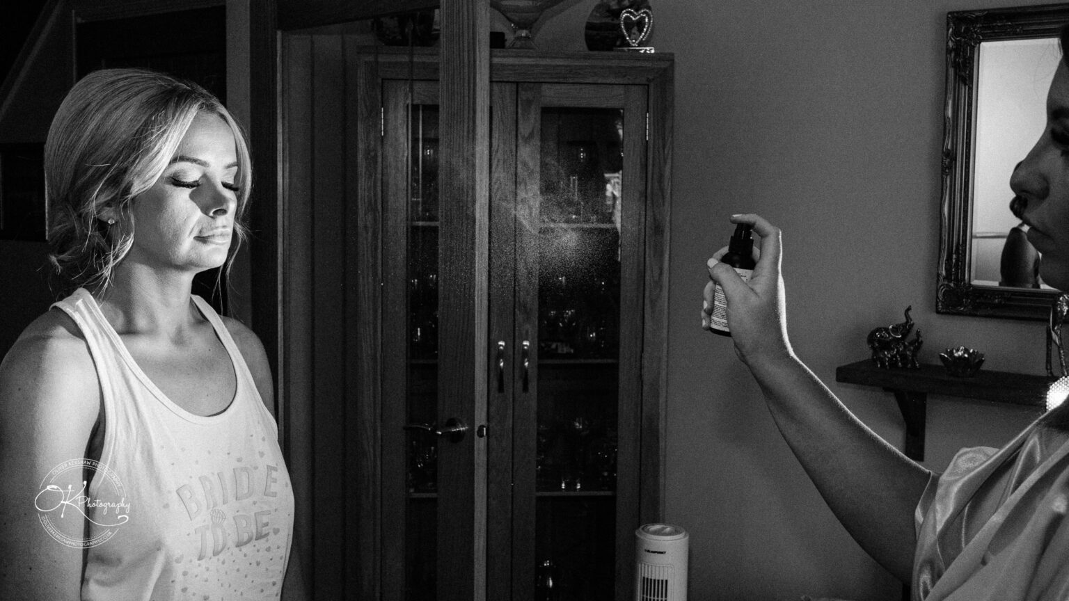 A woman with closed eyes is being sprayed with a mist by another person. She is wearing a "Bride to Be" tank top.