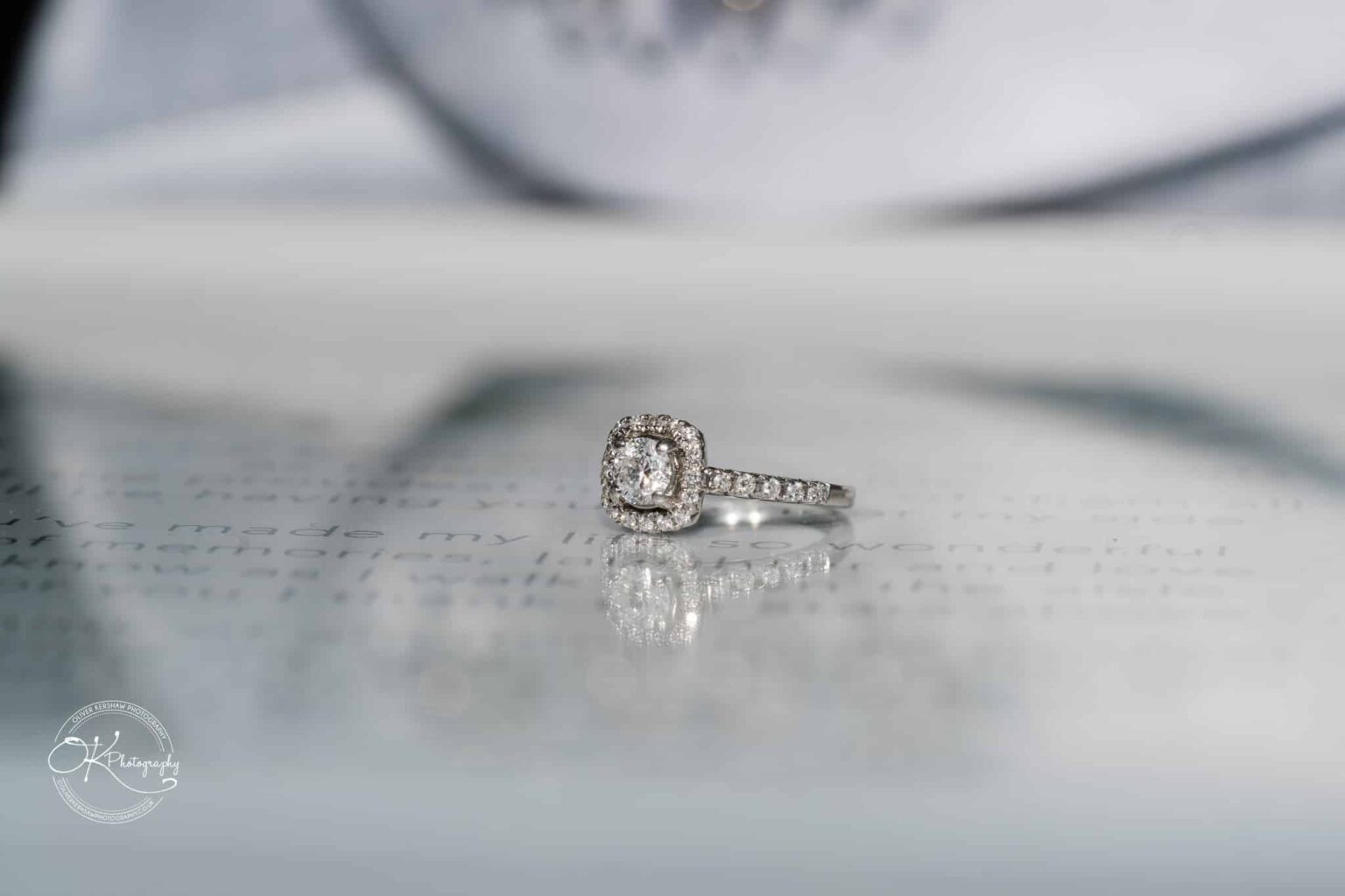 Close-up of a diamond engagement ring with a cushion cut centre stone and a diamond-encrusted band placed on a blurred written surface.
