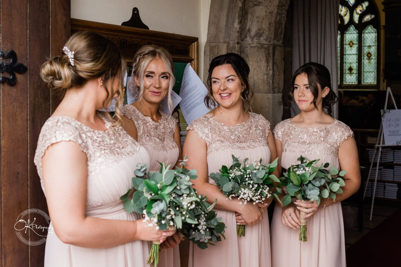 Four bridesmaids in light pink lace dresses, holding green and white bouquets, standing inside a church doorway, smiling and talking.