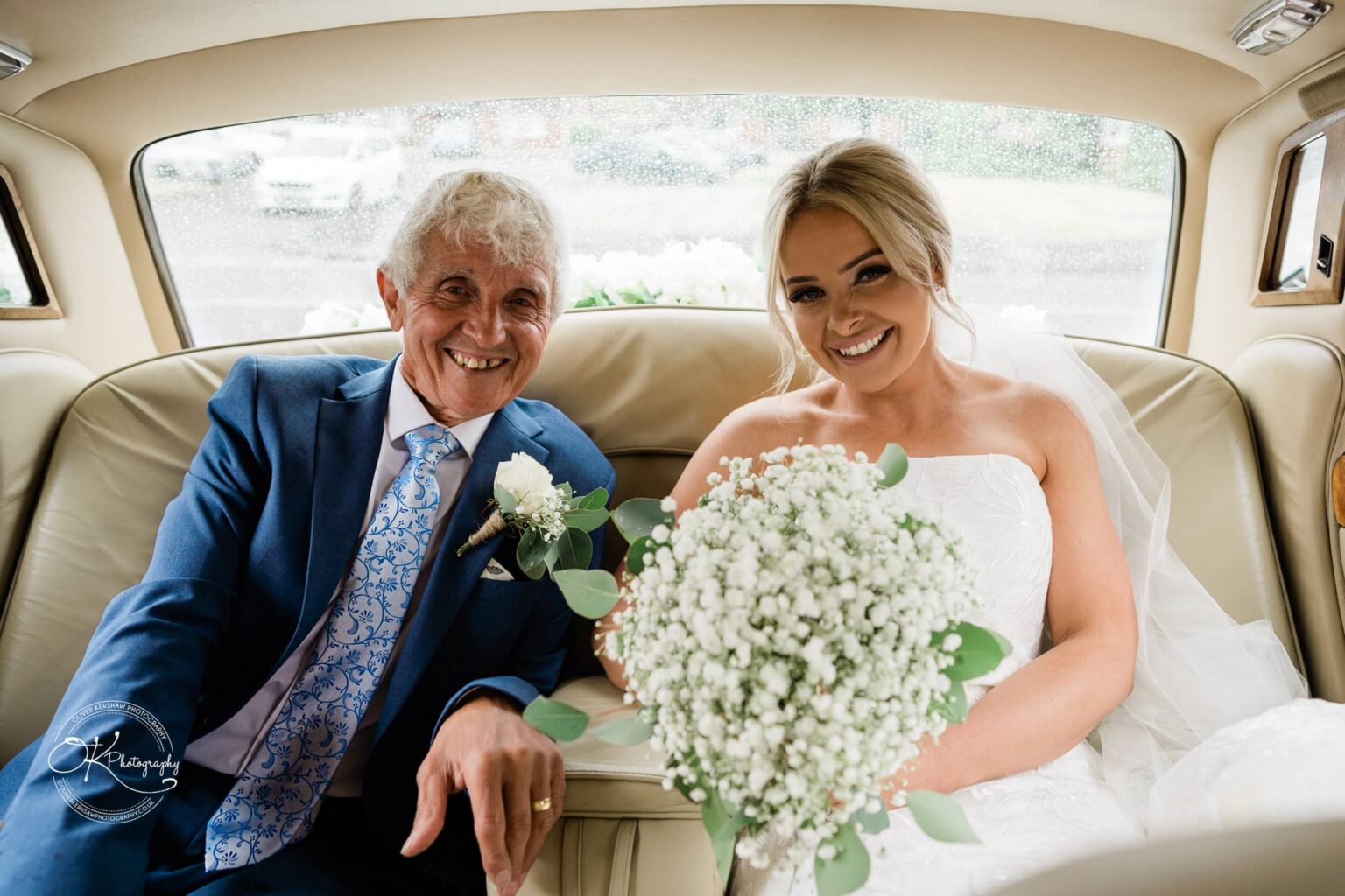 A bride in a white wedding dress holding a bouquet of white flowers sits next to an older man in a blue suit and floral tie inside a car.