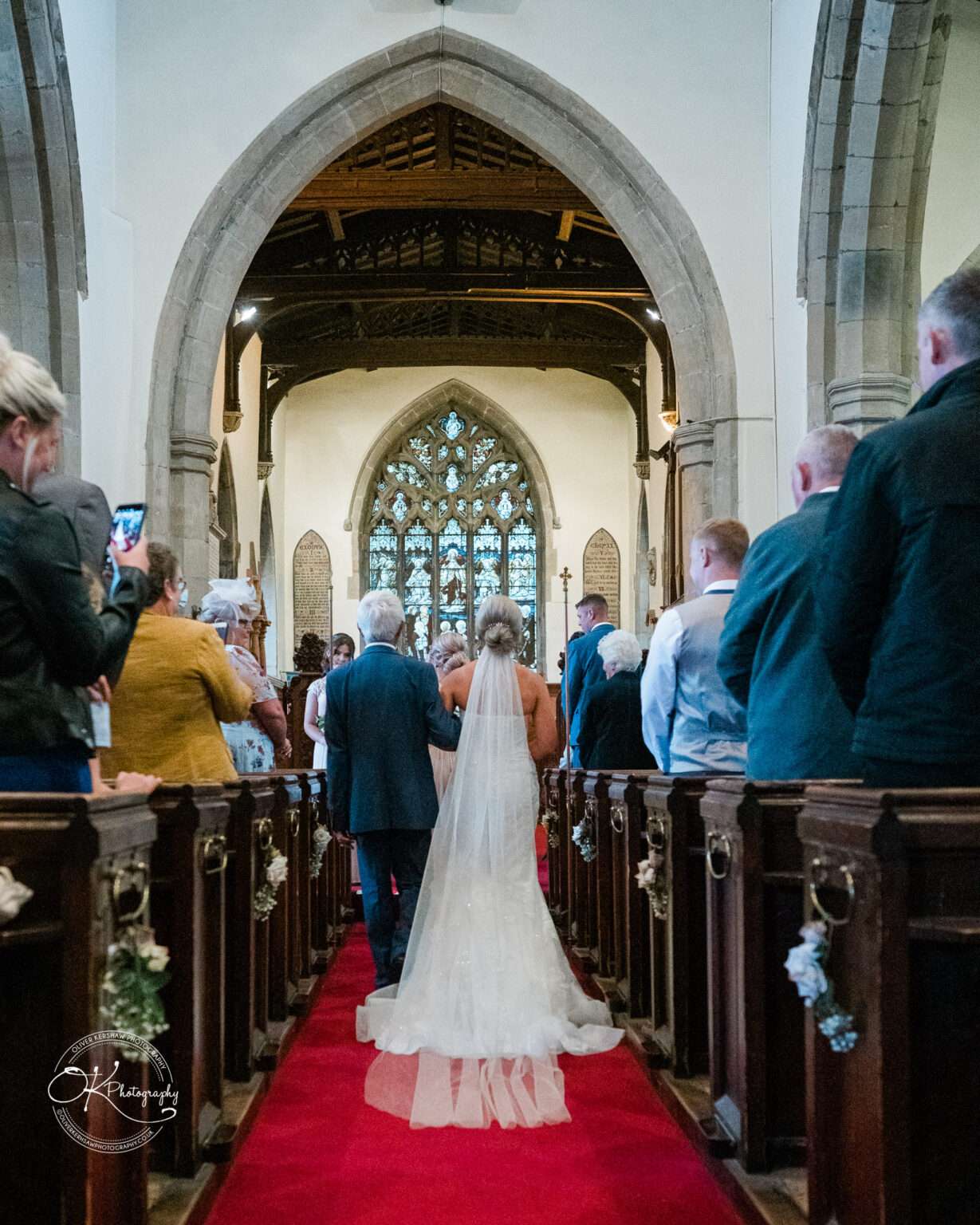 Bride walking down the aisle of a church on a red carpet, accompanied by an older man.