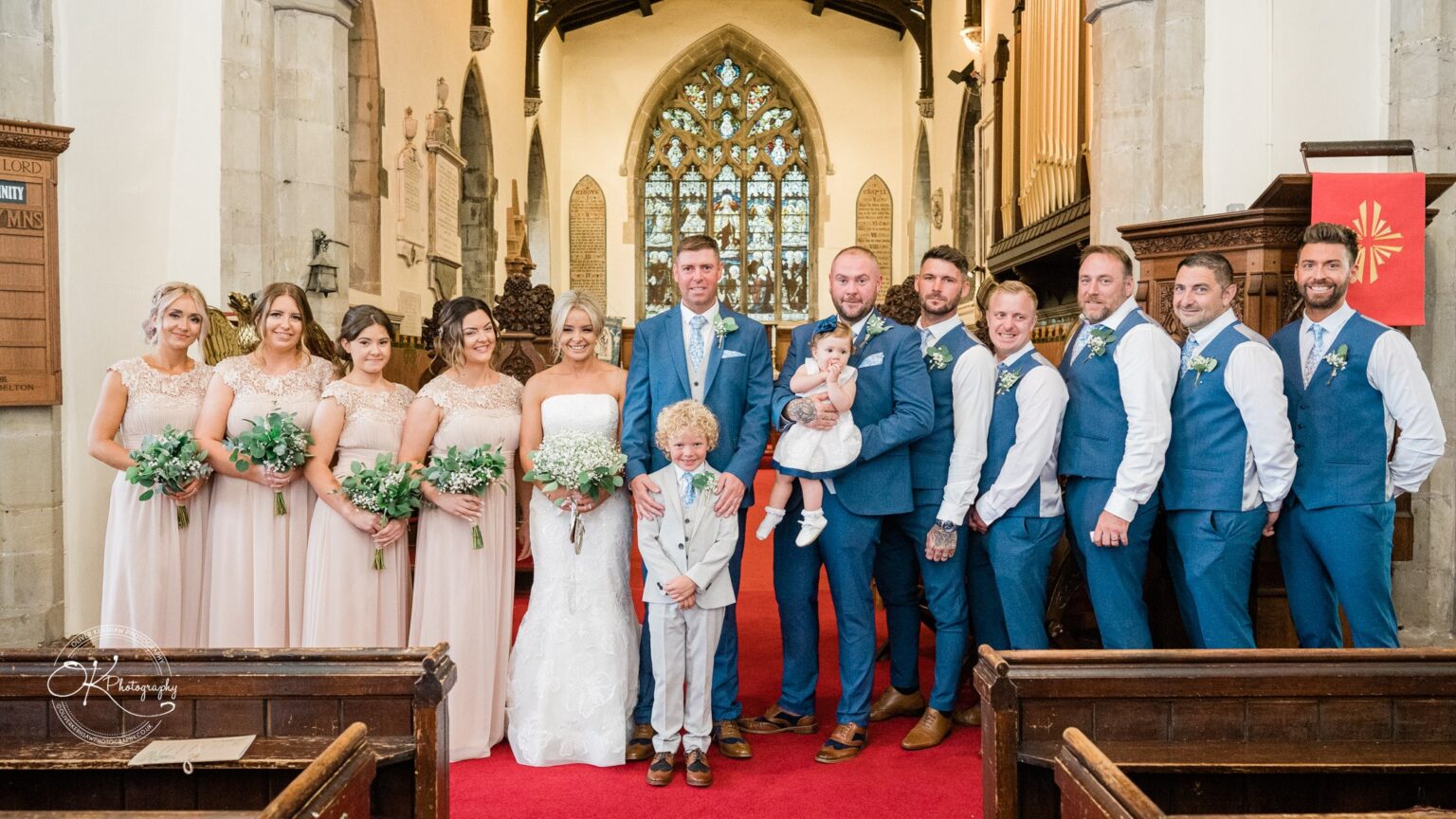A wedding party posing inside a church, with the bride and groom in the center, bridesmaids in pink dresses on the left, and groomsmen in blue suits on the right.