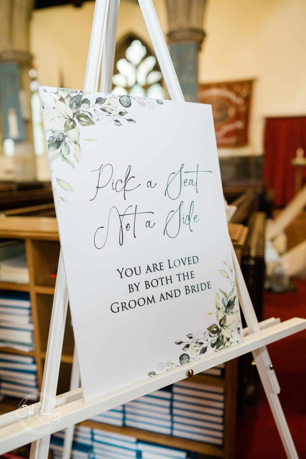 Wedding sign on an easel that reads "Pick a Seat Not a Side. You Are Loved by Both the Groom and Bride," with floral illustrations.