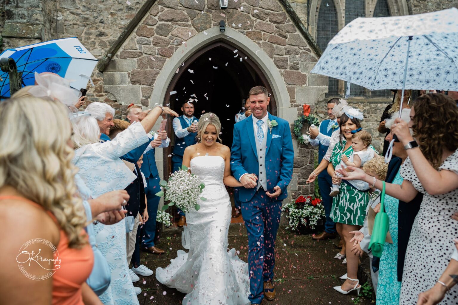 Newly married couple walking out of a stone church while guests celebrate by throwing confetti and holding umbrellas.