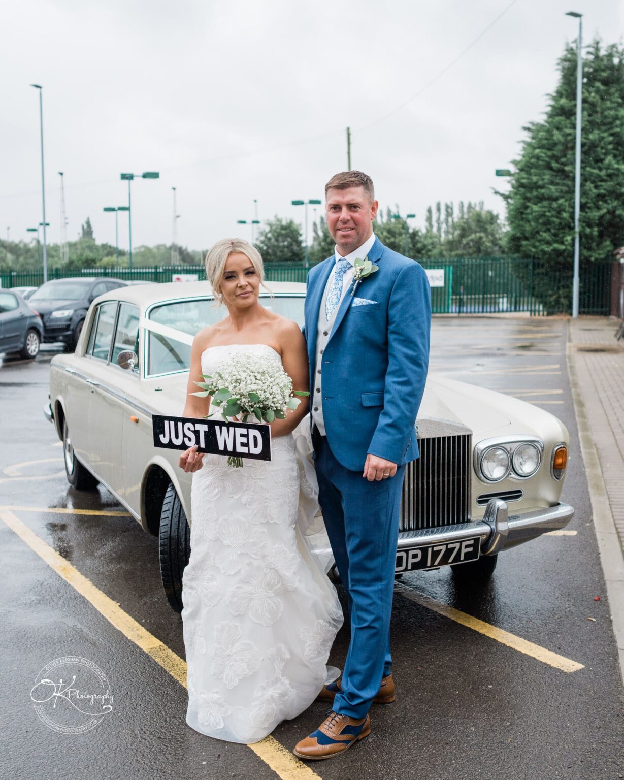 Bride and groom standing in front of a classic car, with the bride holding a "Just Wed" sign.
