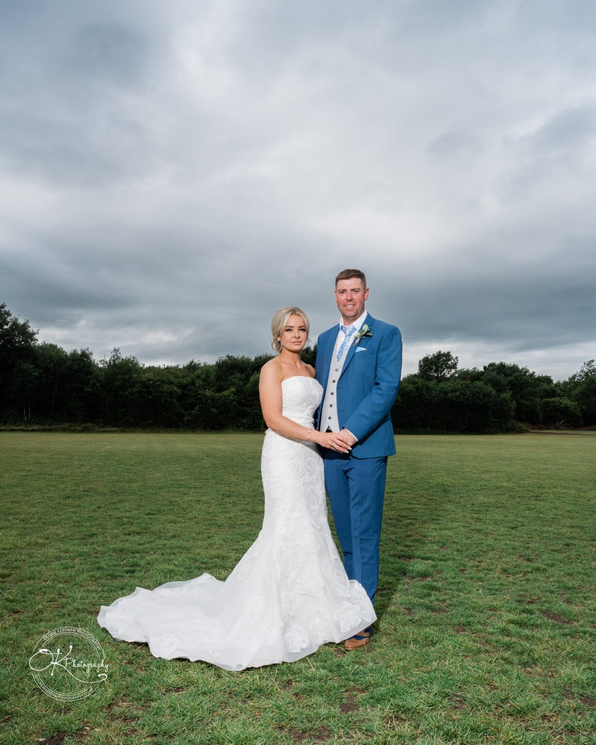Bride and groom holding hands and standing on a grassy field with a cloudy sky in the background.