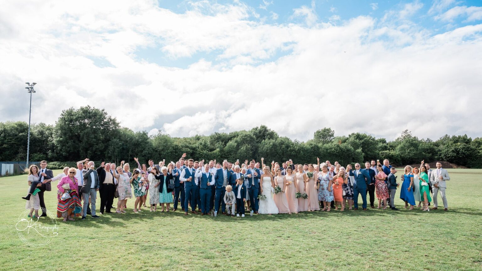 Group of people posing outdoors on a grassy field, with trees and a cloudy sky in the background.