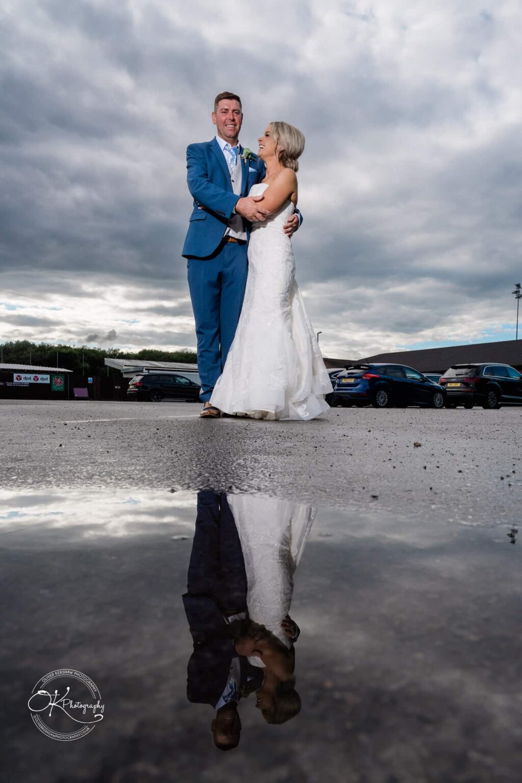 A newlywed couple poses for a photo outdoors, with their reflection captured in a puddle on the ground.
