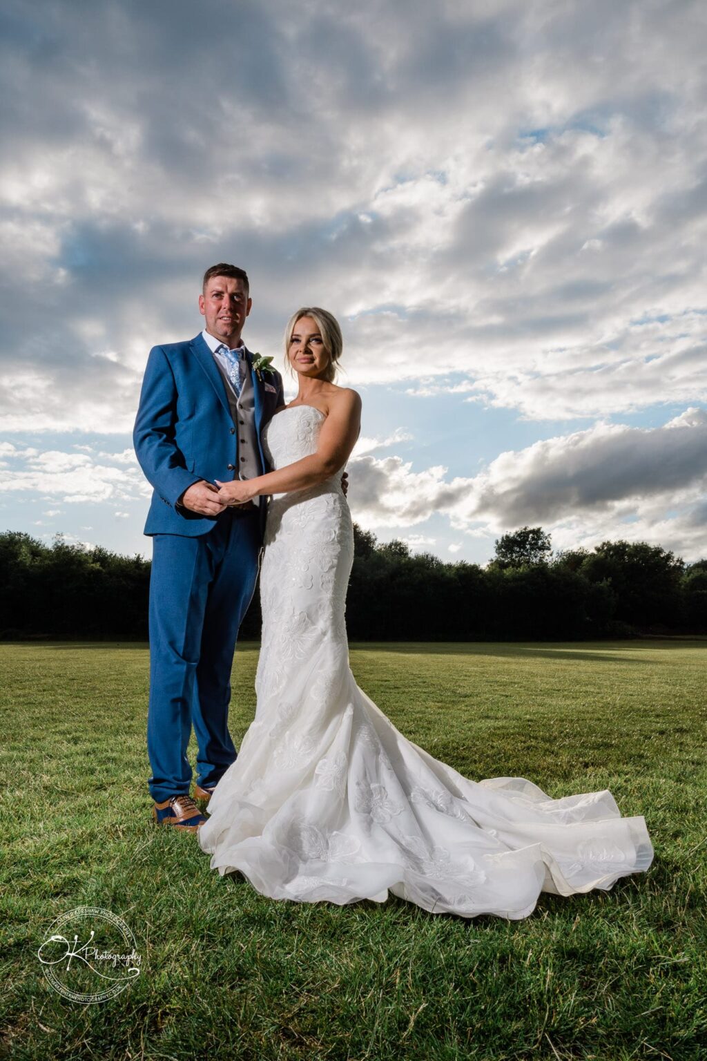 Bride and groom standing on grass, with the bride wearing a white gown and the groom in a blue suit, under a cloudy sky.