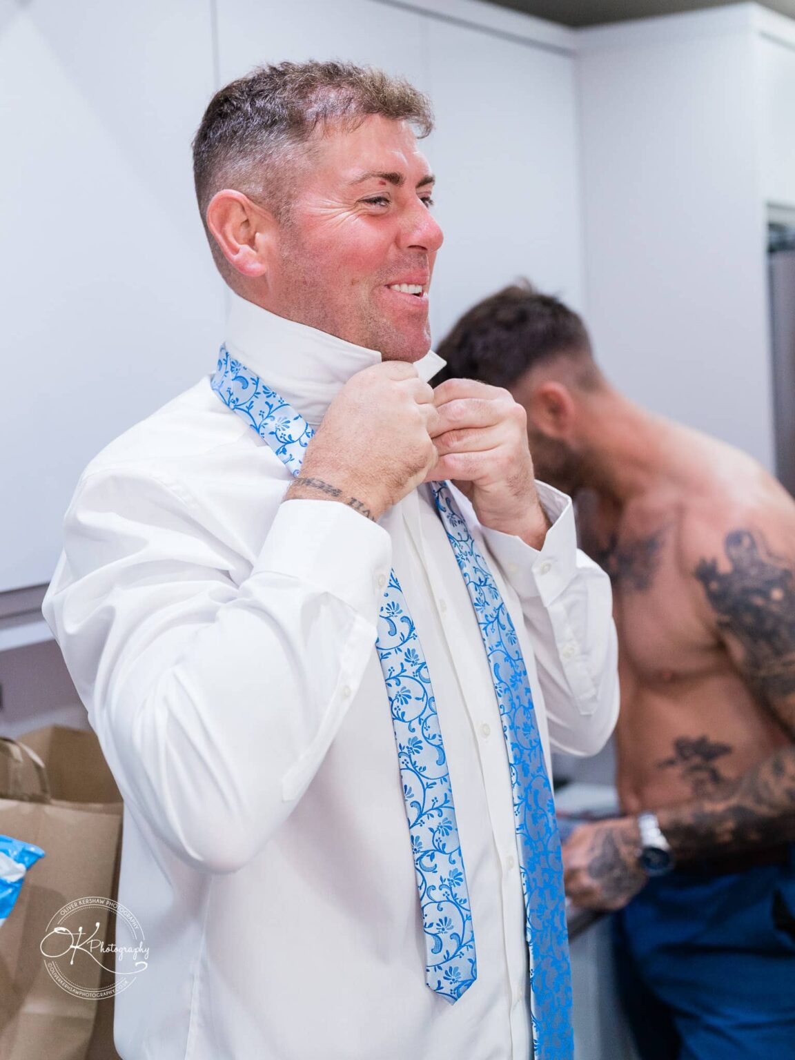 A man adjusting a white shirt and a blue patterned tie with another person in the background, possibly getting dressed.