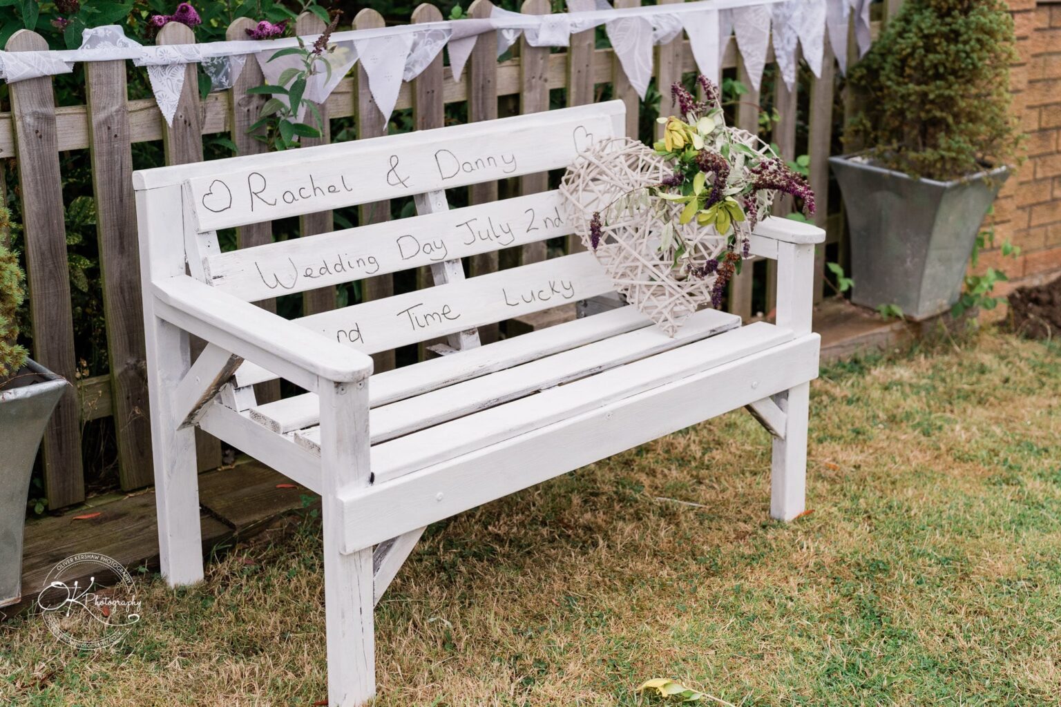A white wooden bench with personalised handwriting reading "Rachael & Danny, Wedding Day July 2nd, Second Time Lucky." A white woven heart with flowers is placed on the bench. In the background, there is a wooden fence adorned with bunting.
