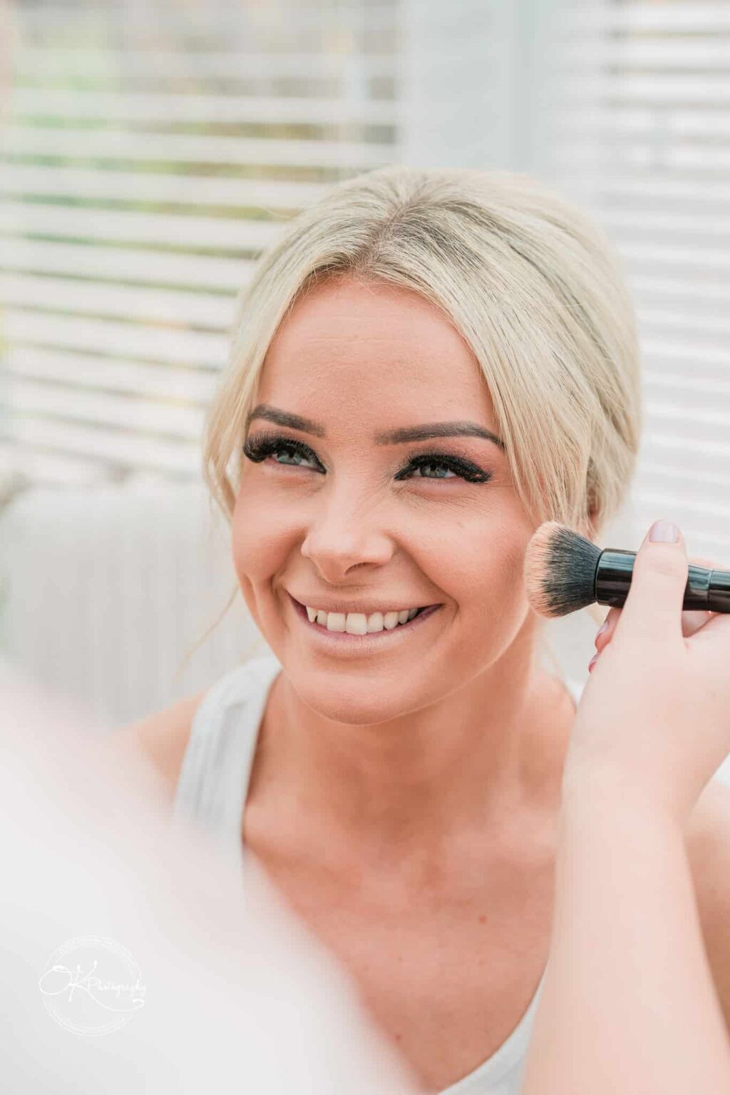 Smiling woman with blonde hair having makeup applied with a brush.