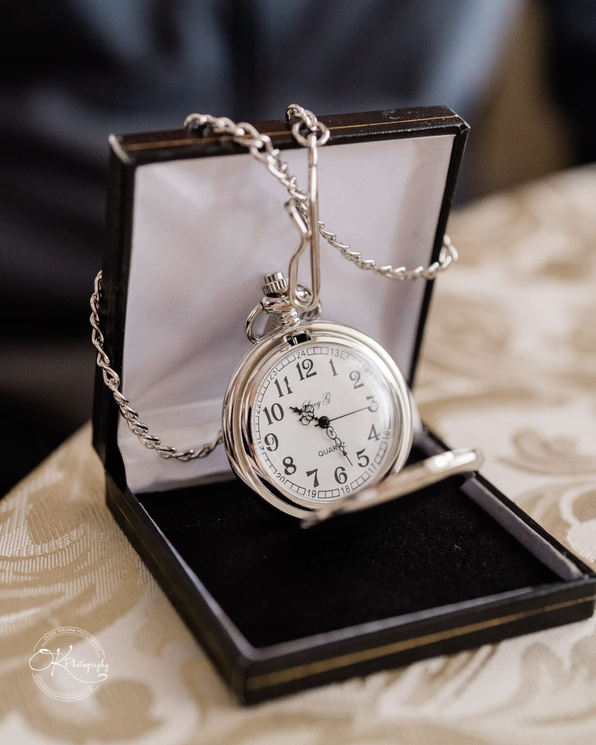 Silver pocket watch with a chain displayed in a black box lined with white fabric.