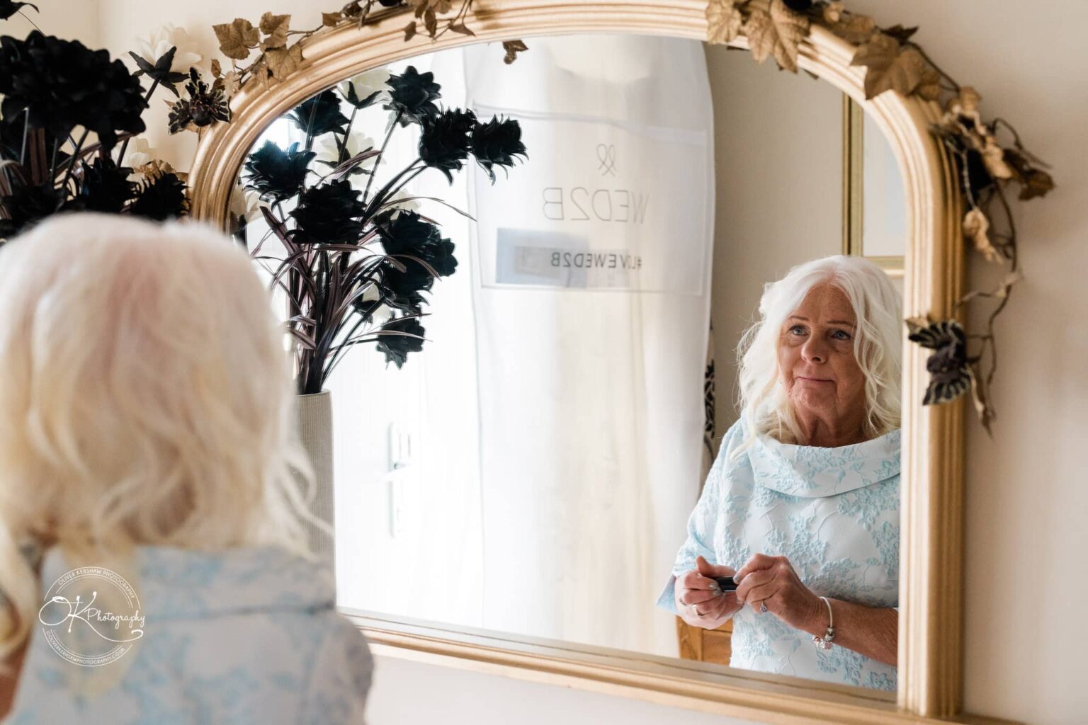 An elderly woman with white hair, wearing a light blue dress, is looking at herself in a mirror while holding a small object.