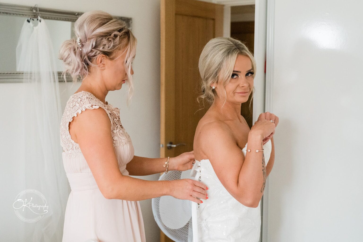 A woman helps another woman adjust her wedding dress in a brightly lit room.