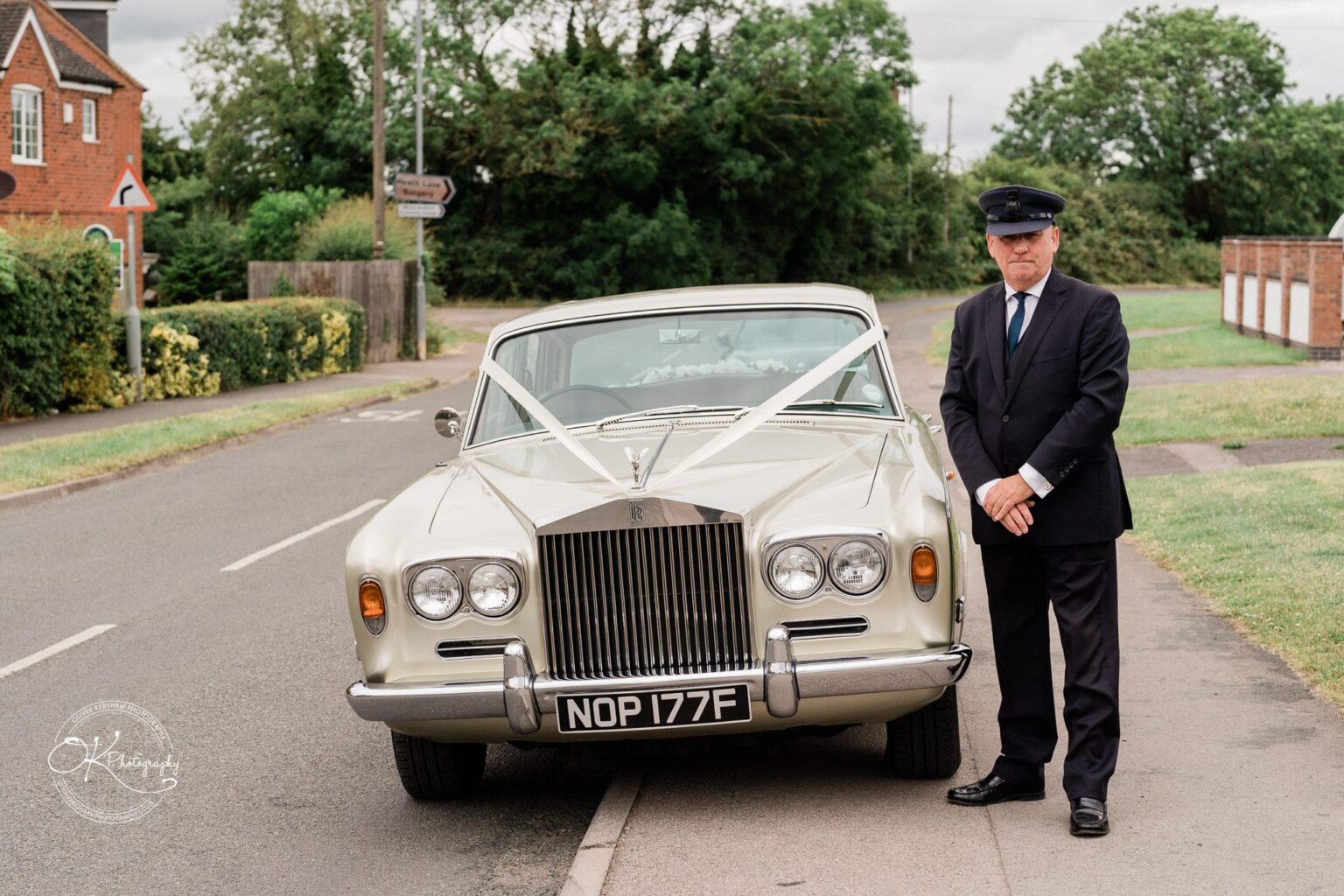 A chauffeur in a suit and cap standing beside a cream-coloured vintage Rolls-Royce decorated with white ribbons.