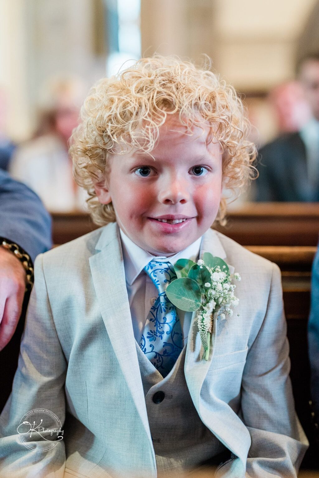 A young boy with curly blonde hair wearing a light grey suit, blue patterned tie, and boutonnière with greenery and white flowers.
