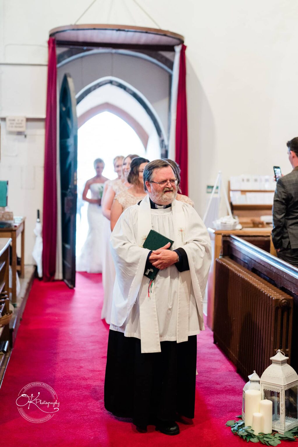 Priest standing in a church aisle with a bridal procession behind him, red carpet and decorative lanterns with candles on the right.