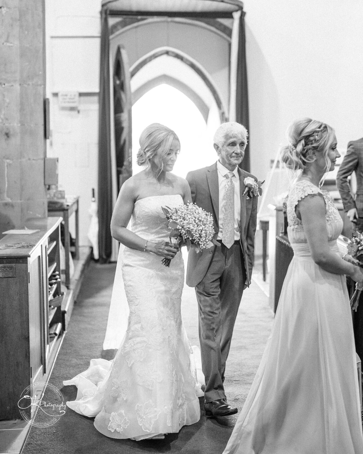 A bride in a strapless wedding gown walks down the aisle with an older man in a suit, holding a bouquet, followed by a bridesmaid in a light-coloured dress inside a church.