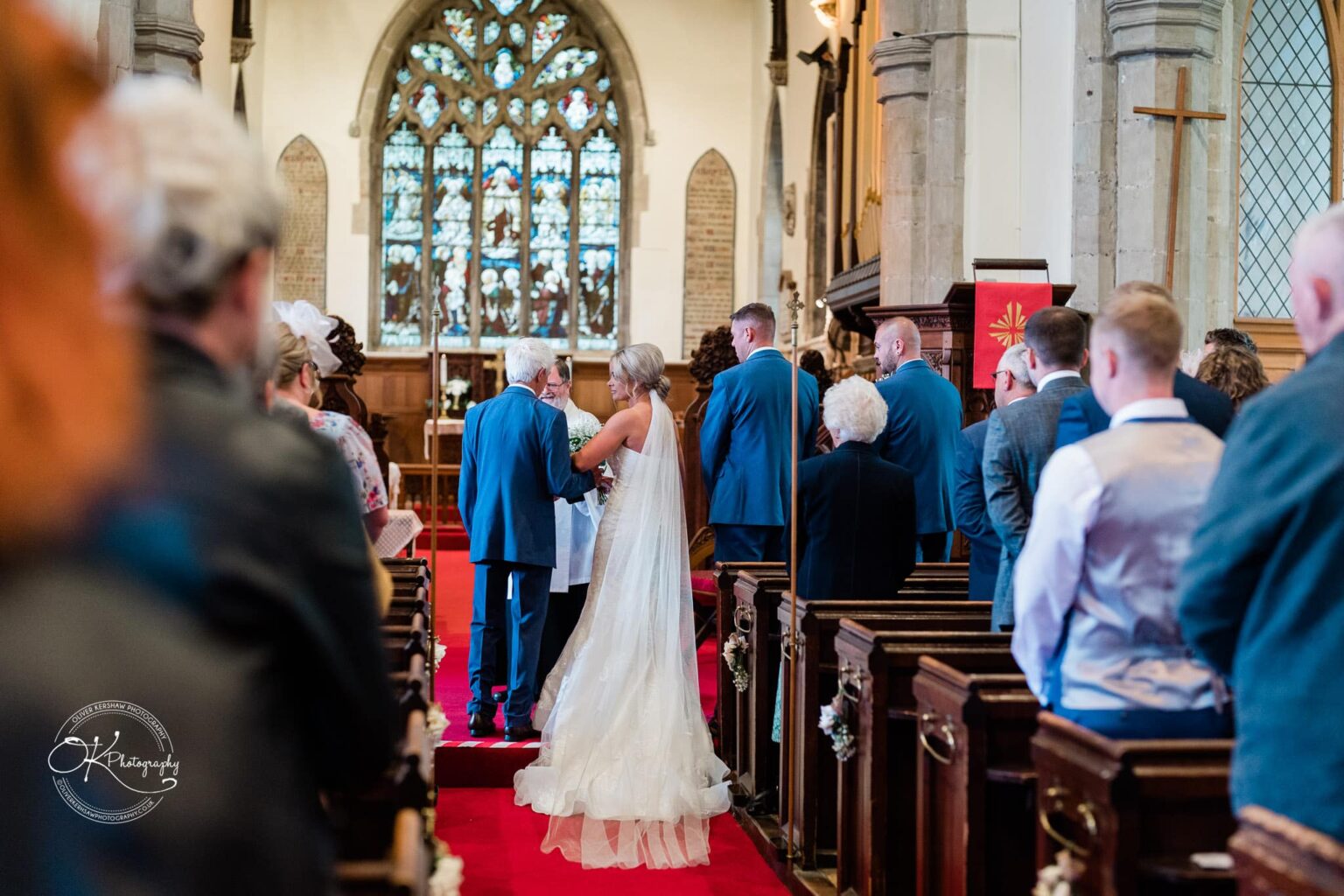 Bride in a white dress and veil walking down the aisle of a church accompanied by an elderly man, with wedding guests standing and watching.