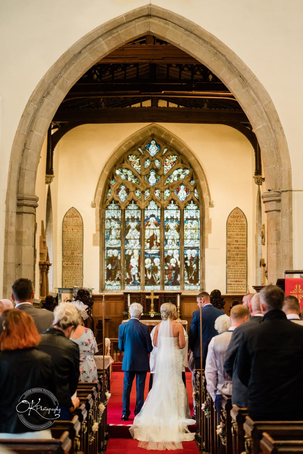 Bride in white gown and veil walks down the aisle inside a church with gothic architecture, stained glass windows, and guests standing on either side.