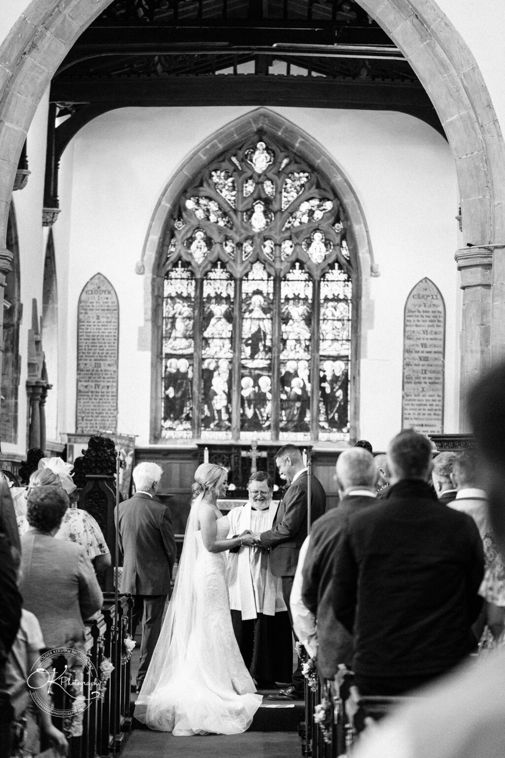 Bride and groom exchanging vows in a church ceremony, with guests seated and a priest officiating under a stained glass window.