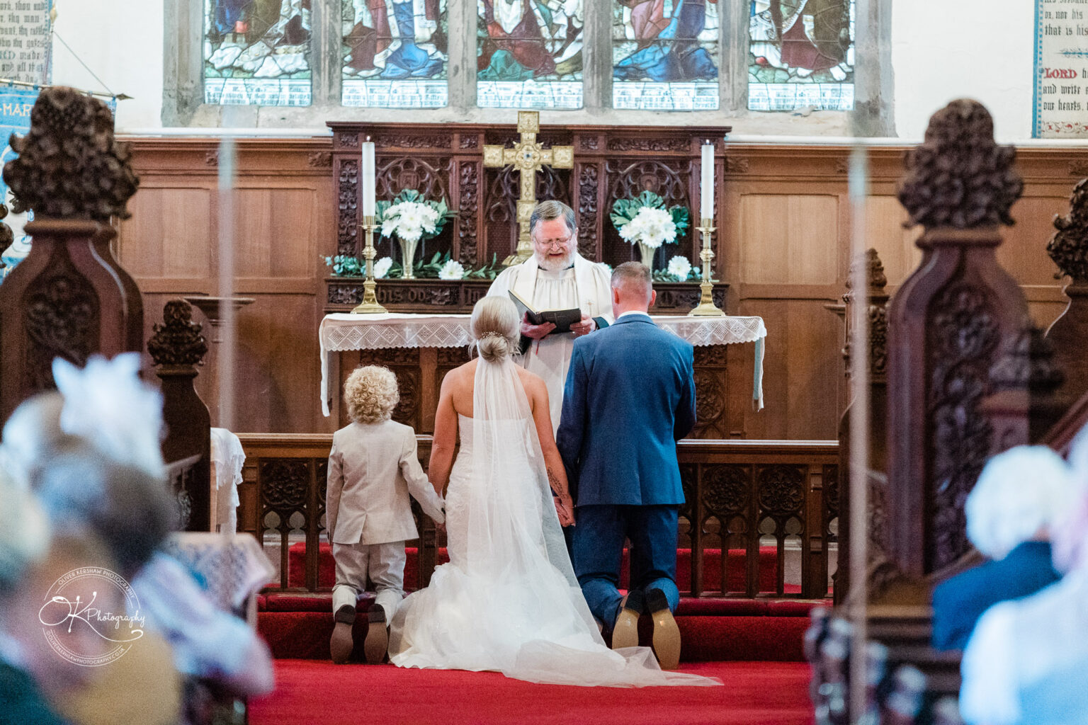 Bride and groom kneeling at the altar with a child, holding hands, during a church wedding ceremony led by a priest.