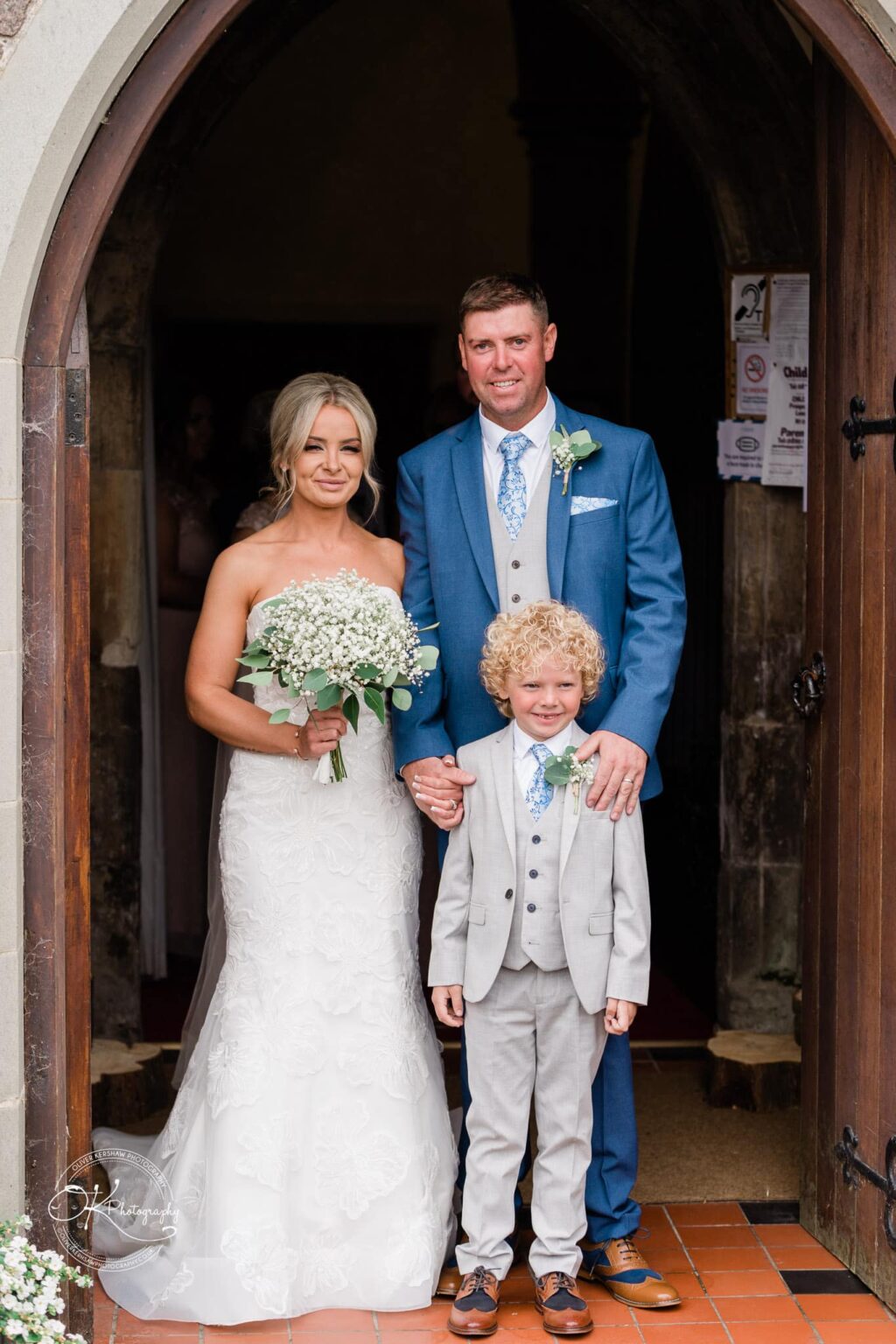 Bride in a white dress holding a bouquet, groom in a blue suit, and young boy in a light grey suit standing together at a church doorway.